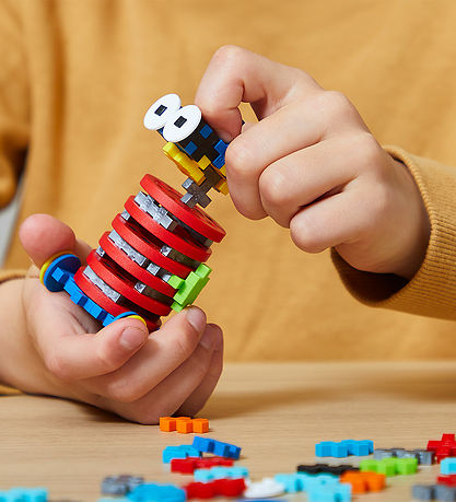 Hands building a toy robot with red, blue, and yellow interlocking cross pieces on a light wooden table.