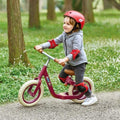 Child in a red helmet and pads riding a red balance bike with white tires and wooden accents on a gravel path.