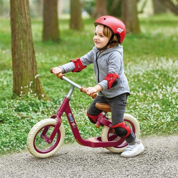 Child in a red helmet and pads riding a red balance bike with white tires and wooden accents on a gravel path.
