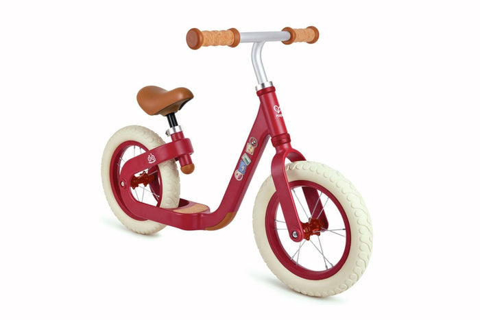 Red balance bike with a curved frame, cream wheels, brown seat, and cork-style handlebar grips.