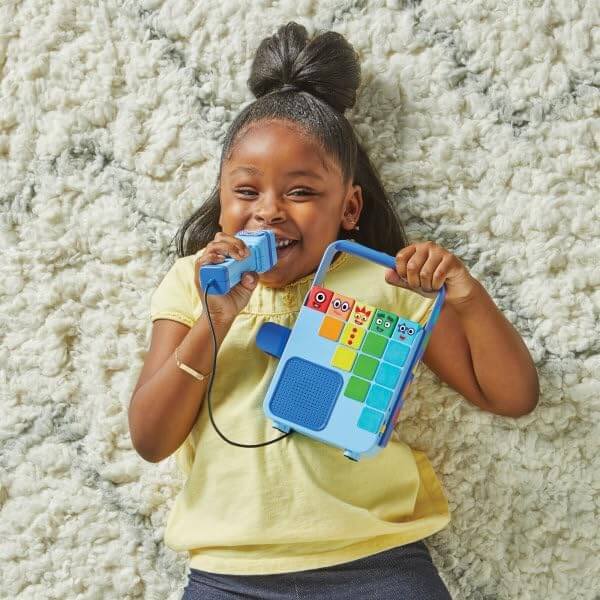 A girl on a rug sings into a blue toy music player with colorful Numberblocks buttons and a wired microphone.