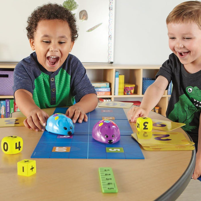 Two boys play with blue and purple robot mice on a blue grid mat with yellow number dice and number cards.