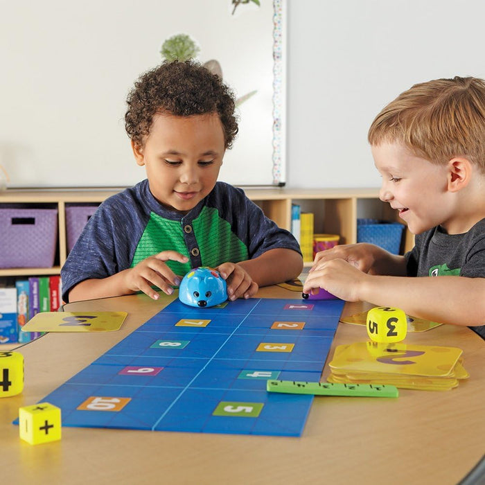 Two boys play with a blue robot mouse on a numbered grid mat with yellow math dice and cards.