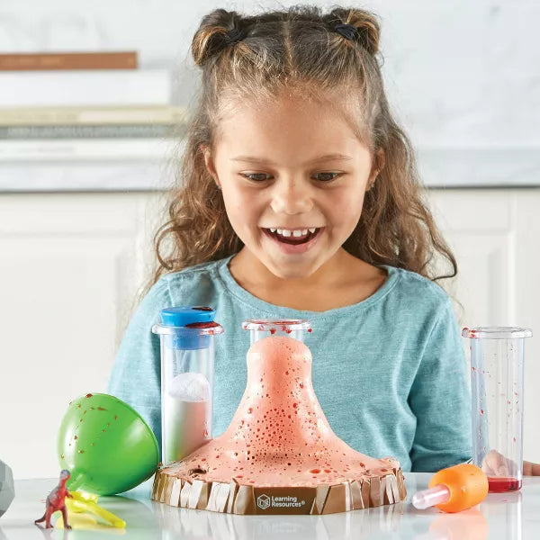 Smiling girl watches a brown toy volcano erupt pink foam, with test tubes, a green funnel, and a red dinosaur.