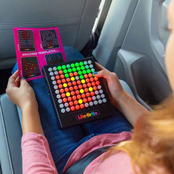 A person in a car holds a square light-up toy with a red strawberry design next to a pink template guide card.