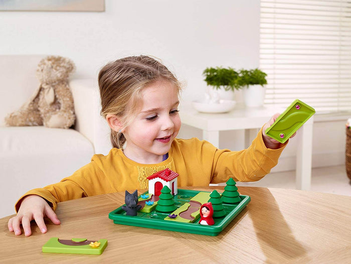 Child playing a forest puzzle with a red hooded figure, wolf, trees, and white house on a green plastic tray.