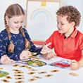 A boy and girl play a math board game with character boards and small cards on a white table.