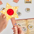 Hands holding a yellow star wand with a red lens over game tokens and an open illustrated instruction book.