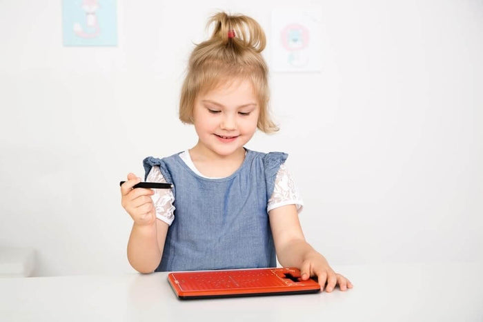 Young girl holding a black magnetic pen over an orange drawing board with a grid pattern on a white table.