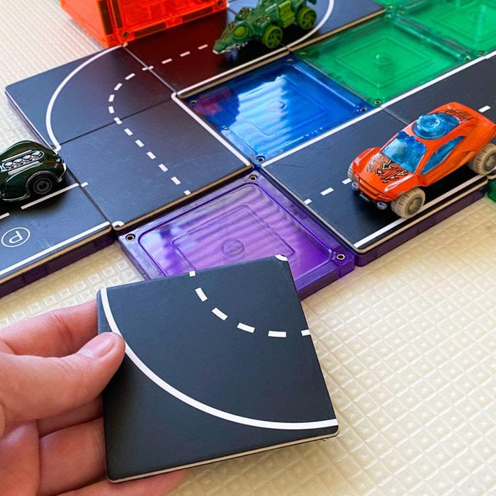 Hand holding a black square road tile topper with white lines over colorful magnetic tiles and toy cars.