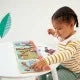 Child in a yellow striped shirt plays with an open magnetic storybook showing a colorful landscape on a white table.