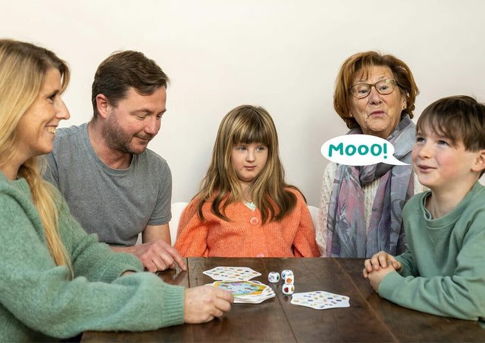 A family plays a card game with hexagonal cards and dice. An older woman says Mooo! in a speech bubble.