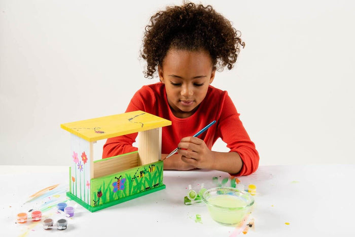 A young girl paints a wooden bird feeder with colorful nature designs using a paintbrush and small paint pots.