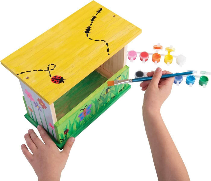 Hands painting a yellow and green wooden bird feeder with bug designs using a paintbrush and small paint pots.
