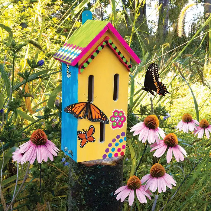 Colorful hand-painted wooden house with narrow vertical slits, set in a garden with pink flowers and butterflies.