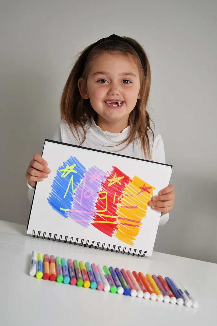 Smiling girl holds a colorful drawing above a row of 25 vibrant markers in various colors on a white table.