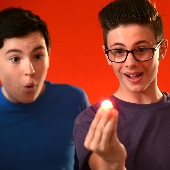 Two boys look amazed as one holds a small glowing orange light between his fingertips against a red background.