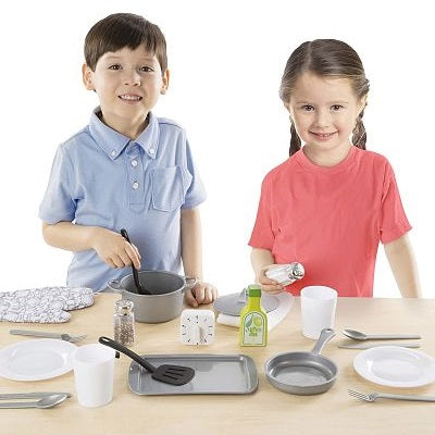 Two children play with a variety of grey and white toy kitchen accessories including pots, pans, and plates.