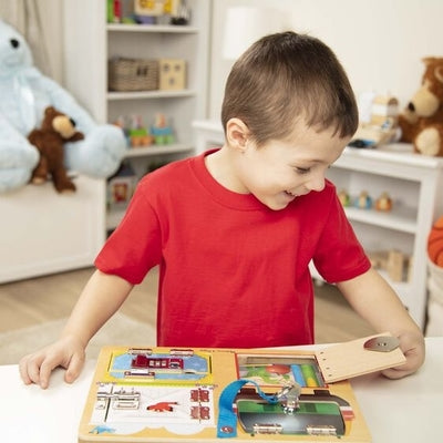A boy in a red shirt smiles while playing with a wooden activity board featuring metal latches and opening doors.