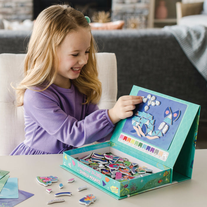 Smiling girl uses sparkly pieces to create a mosaic whale on a teal mermaid-themed activity box with a built-in easel.