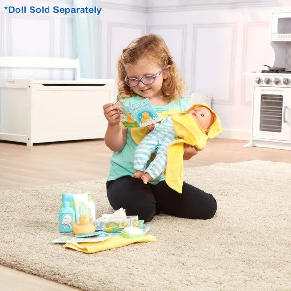 Girl reads a book to a baby doll in a yellow towel. Play bath accessories like a rubber duck are on the rug nearby.