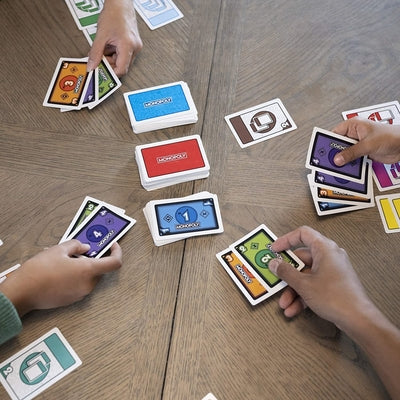 Overhead view of hands playing colorful Monopoly cards with property and action icons on a wooden table.