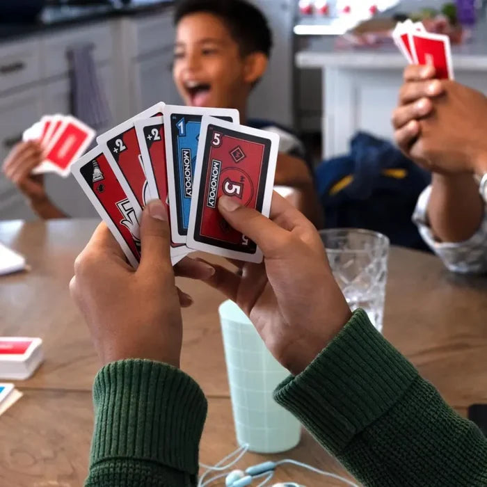 Hands hold a fan of Monopoly Bid cards, including red and blue property and action cards, over a wooden table.