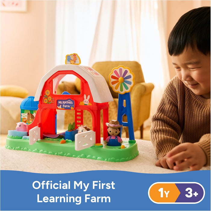 Child plays with a colorful toy farm featuring a red barn, blue windmill, and small animal figures on a green base.