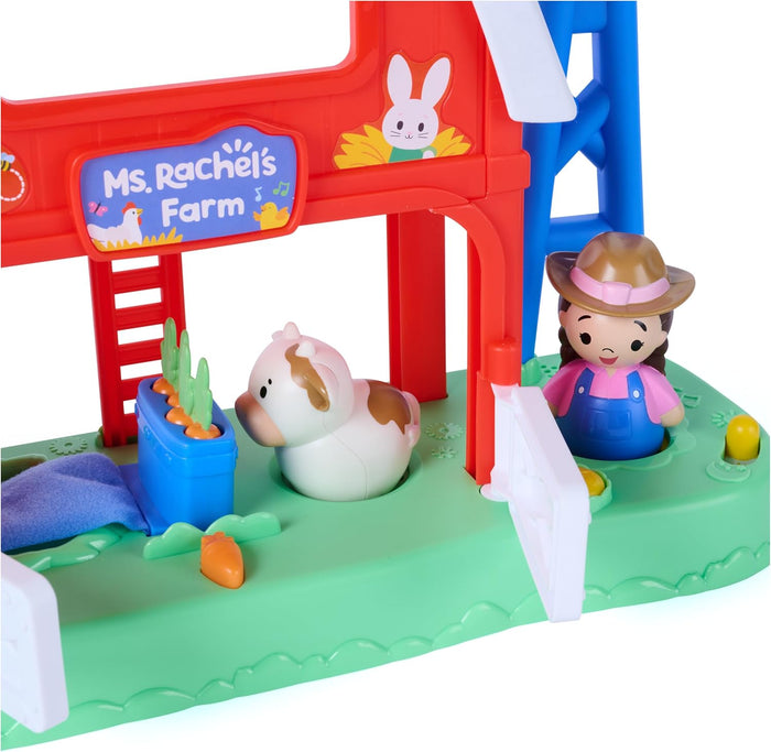 Red and blue toy barn with cow and farmer figures, featuring a vegetable patch and white gate on a green base.