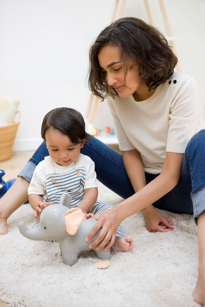 Grey plush elephant toy with pink ears and pull cord, being played with by a mother and toddler on a rug.