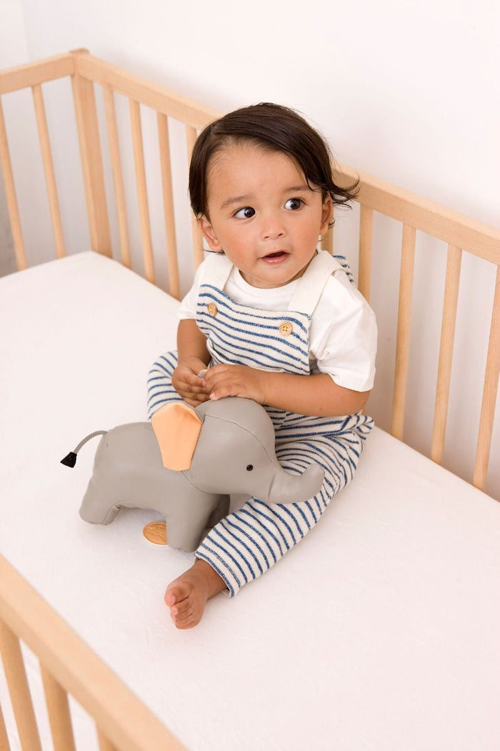 Grey faux-leather toy elephant with peach ears sits in a wooden crib next to a toddler in striped overalls.