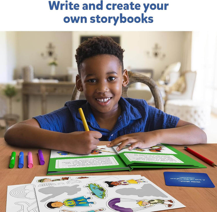 A boy smiles while writing in a green DIY storybook kit with stickers, markers, and prompt cards on a wooden table.