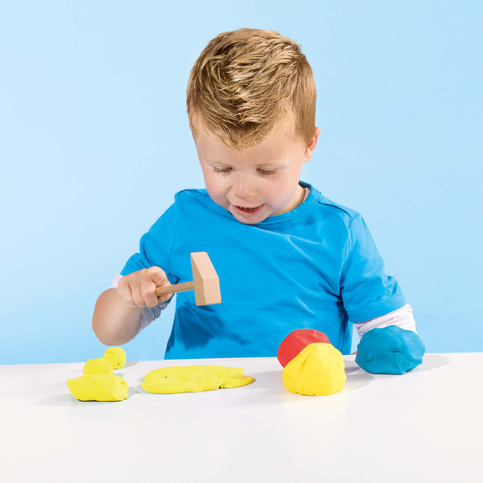 A young boy in a blue shirt uses a wooden hammer to play with yellow, red, and blue dough on a white table.