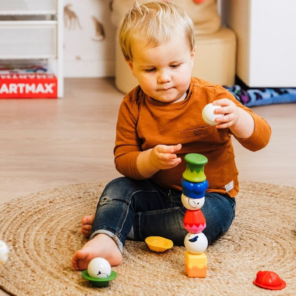 Toddler building a tower with colorful magnetic textured blocks and white spheres with faces on a woven rug.