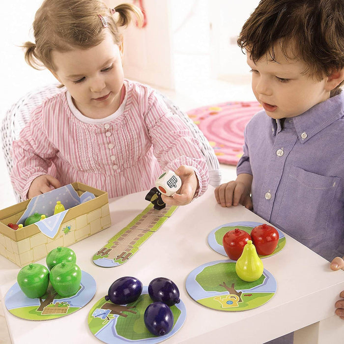 Two toddlers play with chunky wooden fruit, a large white die, and round illustrated cards on a white table.
