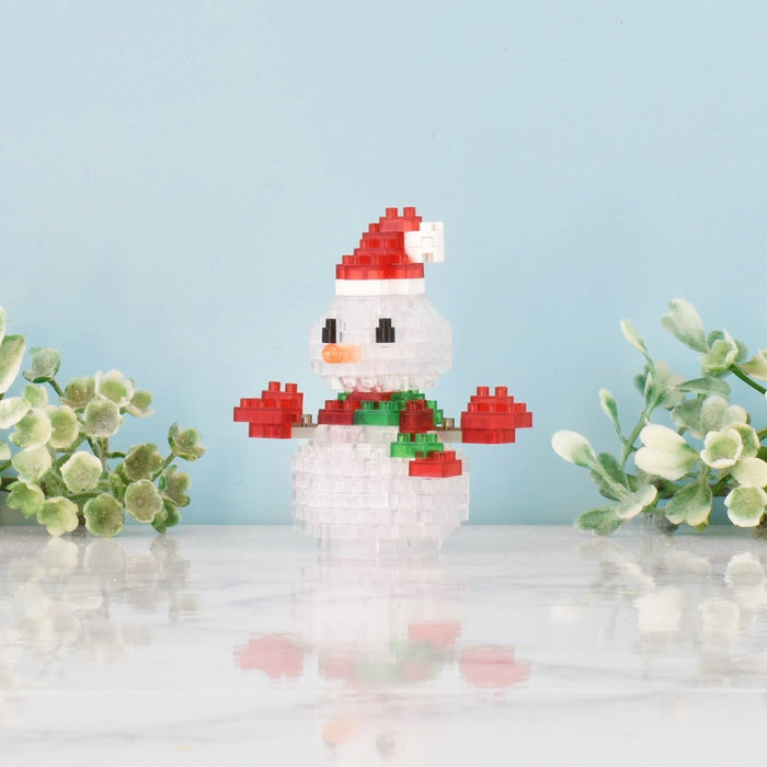 Building block snowman with a red Santa hat, green scarf, and orange nose, standing on a white reflective surface.