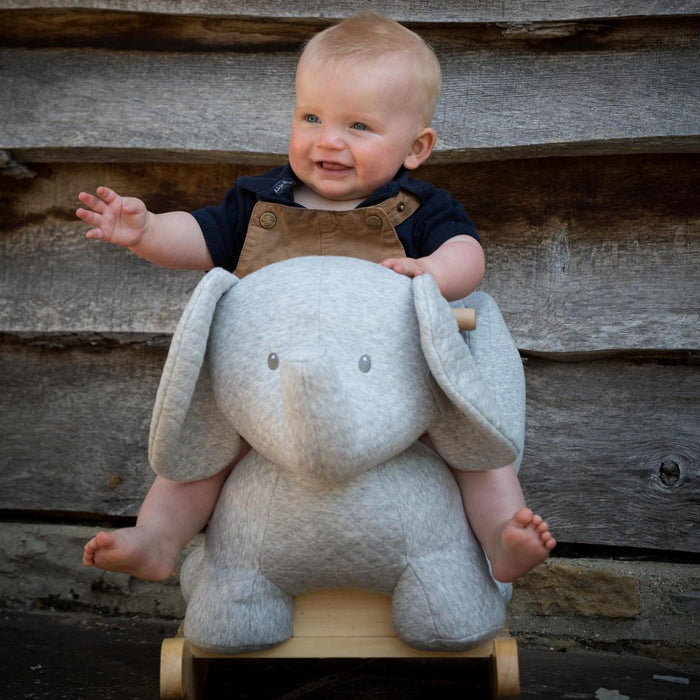 Smiling baby sits on a grey plush elephant rocker with large floppy ears and a wooden rocking base.