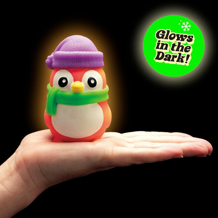 Orange toy penguin with a purple hat and green scarf on an open hand. A green badge reads Glows in the Dark!