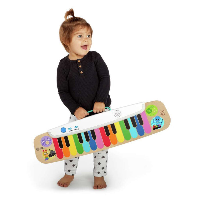 Toddler holding a flat wooden toy keyboard featuring colorful rainbow touch keys and a white handle.