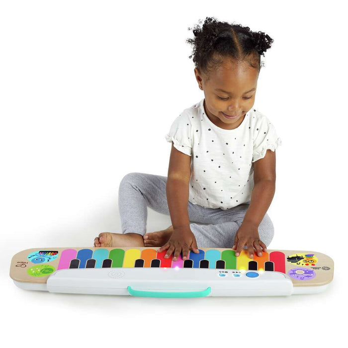 Toddler girl playing with a wooden touch keyboard featuring rainbow-colored keys and a white base with a teal handle.