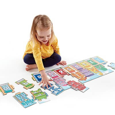 A young girl in a yellow shirt assembling a long, colorful floor puzzle featuring numbered houses.