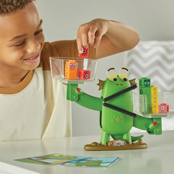 Child adds colorful numbered blocks to a green dragon-shaped balance scale with clear trays.