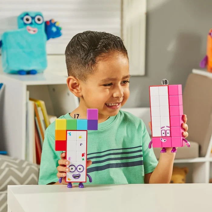 A child holds Numberblock 17, white with paint splatters, and Numberblock 18, a bright pink rectangular toy.