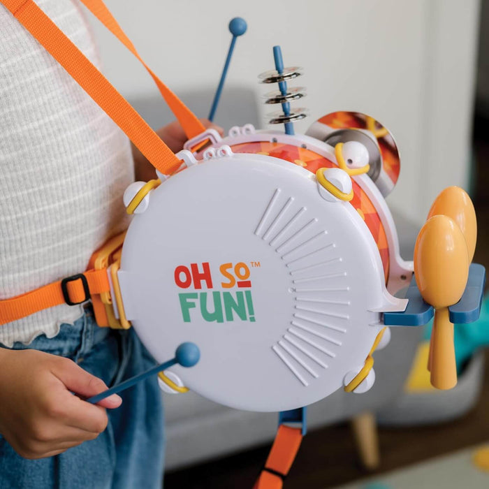 Child wearing a white drum toy with orange straps, yellow maracas, and cymbals, using a blue drumstick.