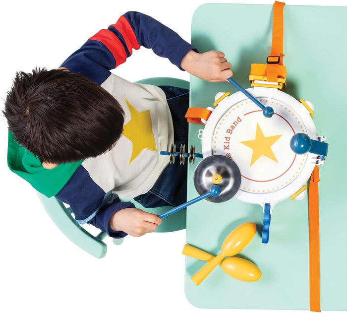 Child playing a white toy drum with a yellow star, blue sticks, a cymbal, and yellow maracas on a light green table.