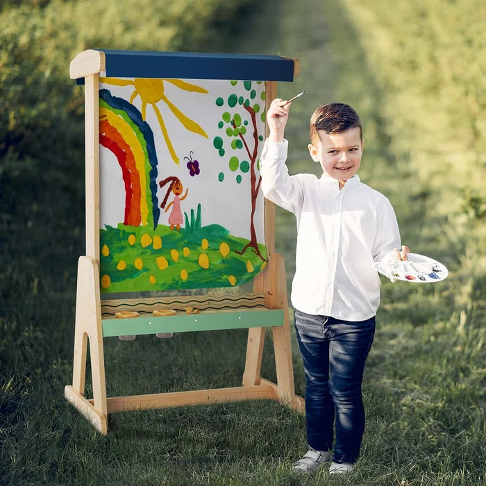 Wooden outdoor easel featuring a colorful rainbow painting, standing in a grassy field next to a boy holding a palette.