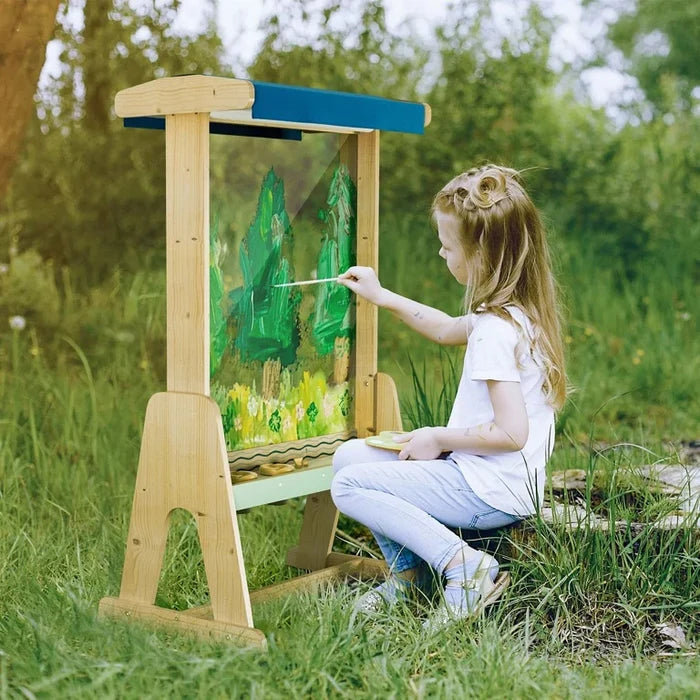 Young girl painting on a wooden outdoor easel with a clear acrylic panel in a grassy yard.