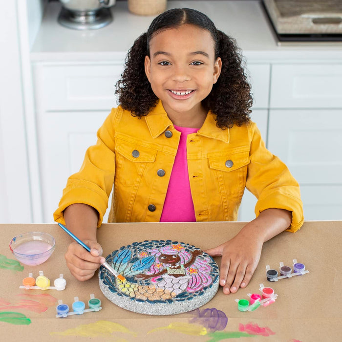 Child painting a round cement stepping stone with a mermaid design using small colorful paint pots.