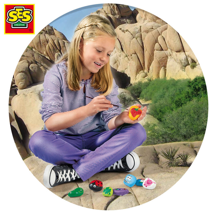 Girl painting a stone to look like a strawberry, with colorful ladybug, sheep, and clover rocks on the ground.