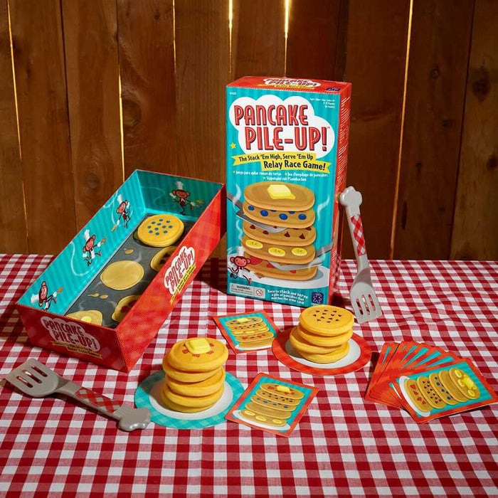 Toy pancakes with toppings, spatulas, and cards on a red checkered tablecloth next to the teal and red game box.